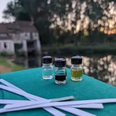 Trois flacons d'huiles essentielles et mouilleurs posés sur un carnet teal, au bord de l'eau devant le Moulin d'Andé au coucher du soleil